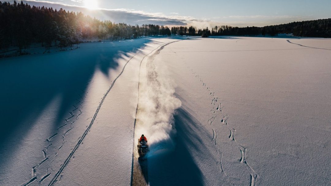 En person kjører snøscooter langs en sti i et vidstrakt snølandskap, og passerer områder perfekt for skøyterentusiaster. Solen står lavt på himmelen og kaster lange skygger over snøen.