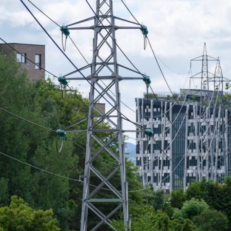 Elektriske kraftoverføringstårn med kabler står høyt i et grønt byområde, og legemliggjør energifleksibilitet. De er omgitt av trær og moderne bygninger under en overskyet himmel, og fremhever harmonien mellom natur og innovasjon.