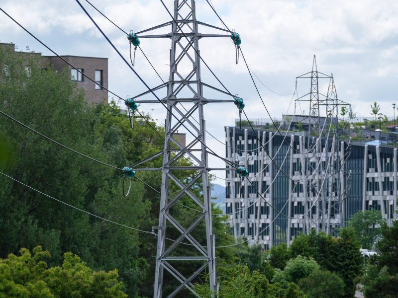 Elektriske kraftoverføringstårn med kabler står høyt i et grønt byområde, og legemliggjør energifleksibilitet. De er omgitt av trær og moderne bygninger under en overskyet himmel, og fremhever harmonien mellom natur og innovasjon.