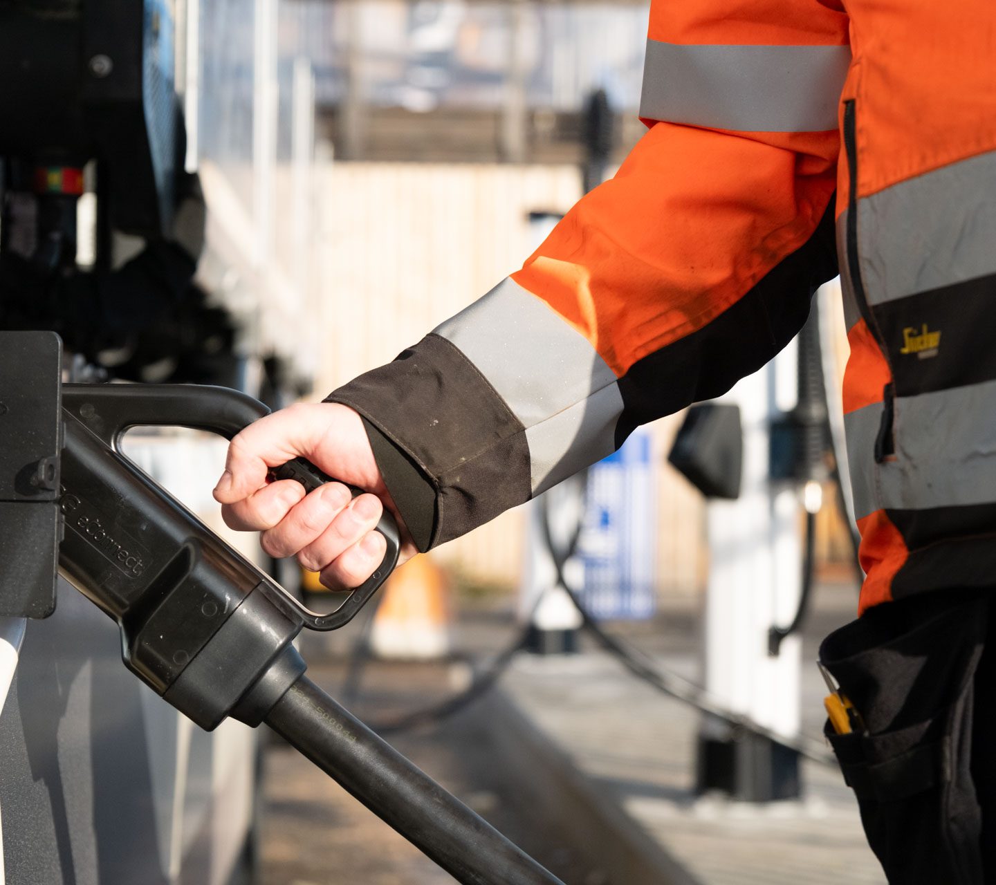 Person i en oransje sikkerhetsjakke som holder en ladeplugg for elektrisk kjøretøy, forbereder seg på å lade et kjøretøy.