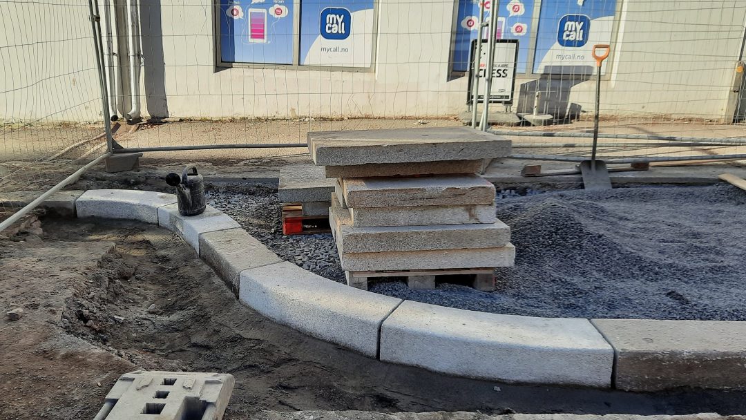 Byggeplassen har stablede betongplater og en buet rad med Stein-lagte steiner på en grusflate, med en vannkanne av plast like ved. Bygningsvinduer er synlige i bakgrunnen.