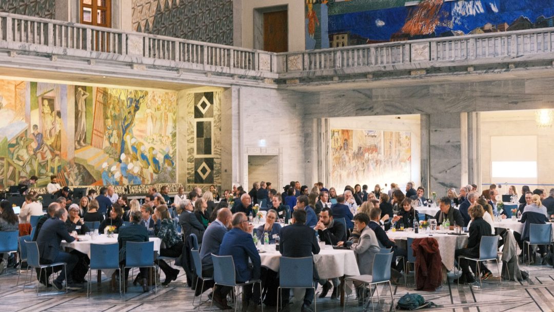 Folk satt ved runde bord i en stor, utsmykket hall med fargerike veggmalerier på veggene.