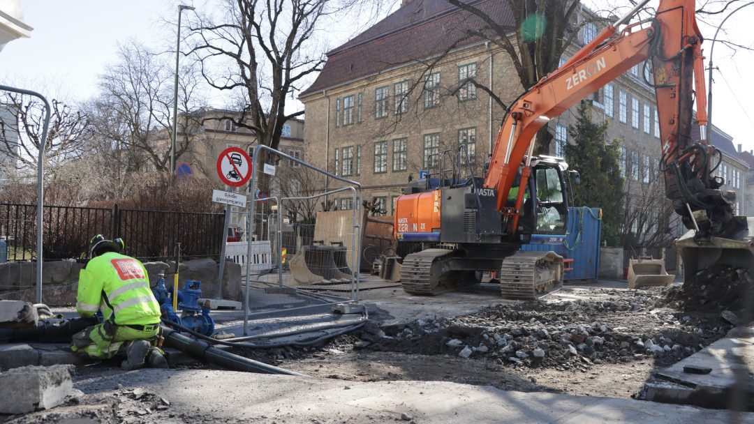 Emission-free machines: An electric excavator in a city street. A man in work clothes is kneeling beside a hole in the ground. Utslippsfrie maskiner: Elektrisk gravemaskin i en bygate. Mann i arbeidstøy sitter på huk ved en hull i bakken