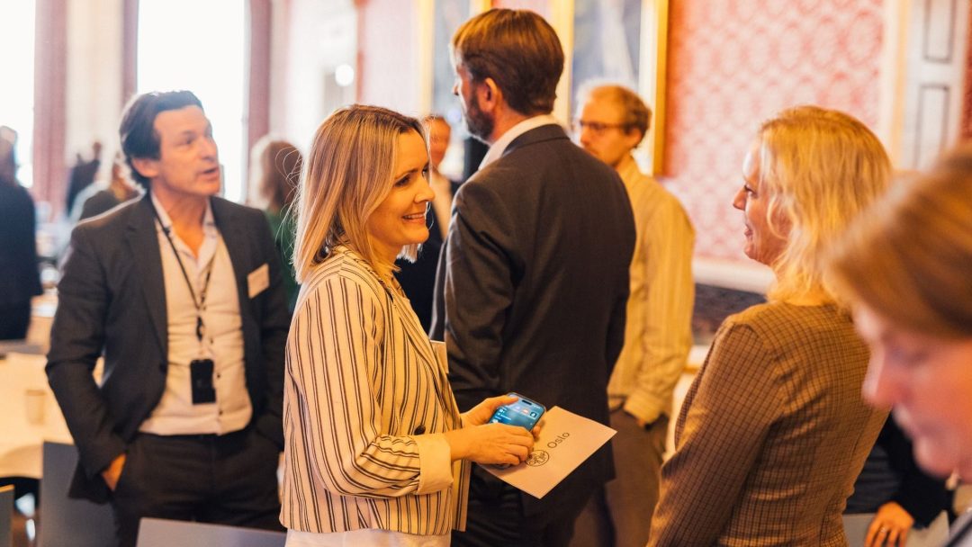Folk engasjerte seg i samtale på et sosialt arrangement i et rom med mønstrede vegger og runde bord. En smilende kvinne holder en drikkeboks og et brettet papir.