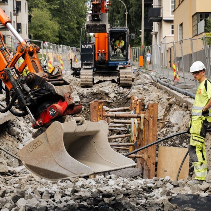 Gravearebider i en bygate. Mann i gult arbeidstøy står ved en gravemaskin, foran et hull i bakken.