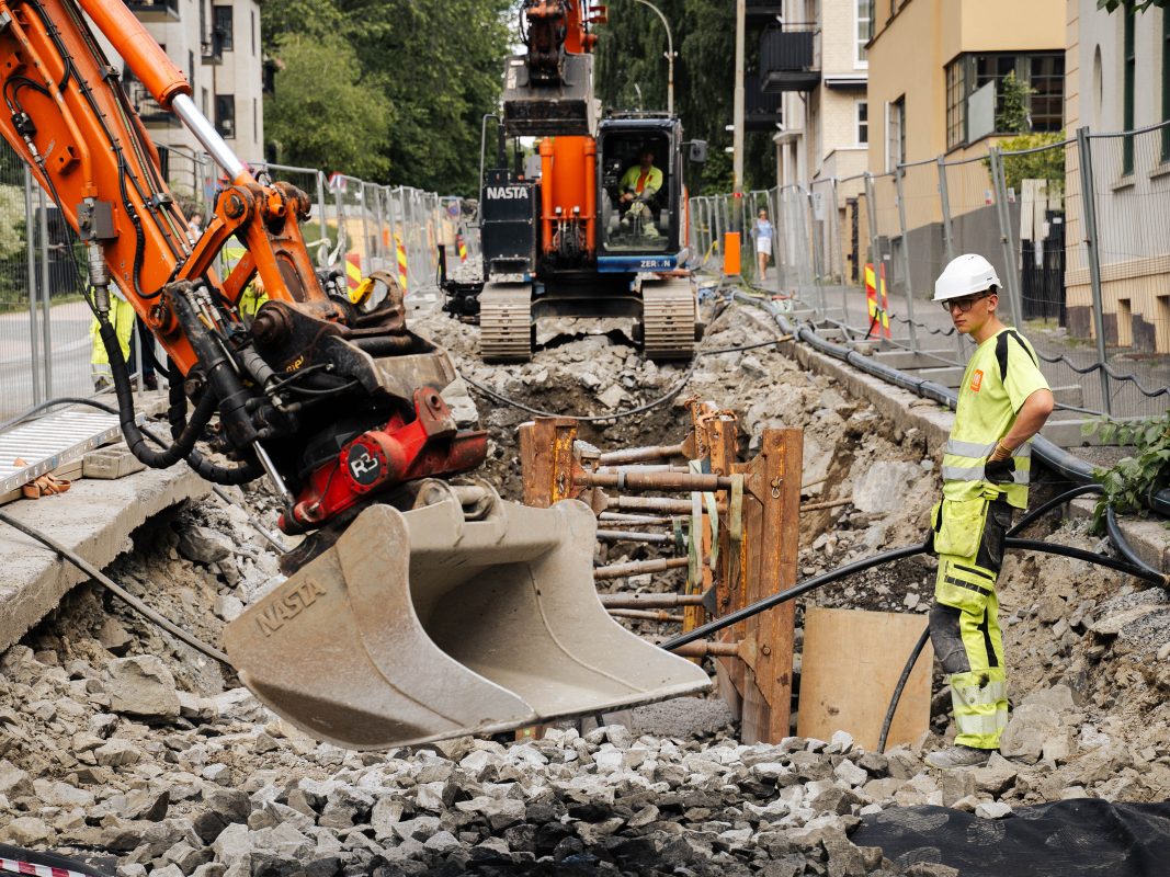Gravearebider i en bygate. Mann i gult arbeidstøy står ved en gravemaskin, foran et hull i bakken.