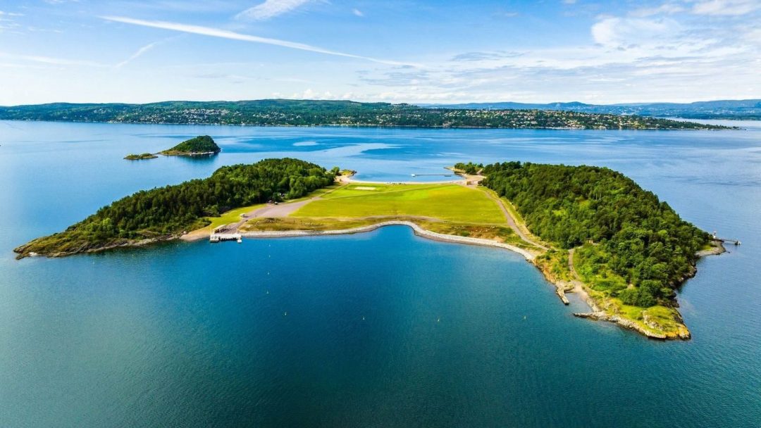 En luftfoto viser en øy utsmykket med grønne felt og trær, omgitt av blått vann under en delvis overskyet himmel. Ideelle badeplasser ligger langs den rolige strandlinjen, og inviterer til avslapning og utforskning.