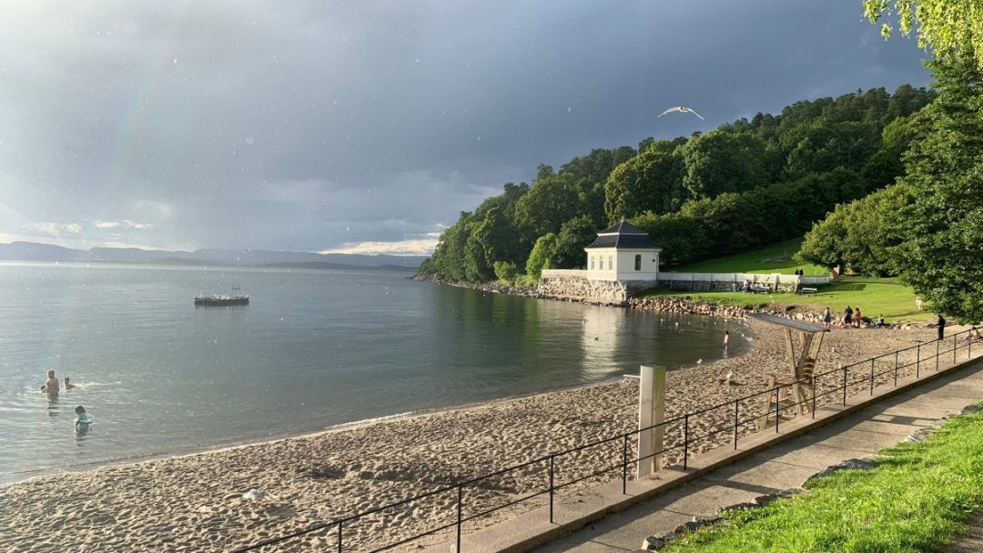 En sandstrand buer seg langs rolig vann ved en av de populære badeplassene, med svømmere, en hvit bygning ved kysten, grønne trær og folk samlet på en gresskledd ås under en delvis overskyet himmel.