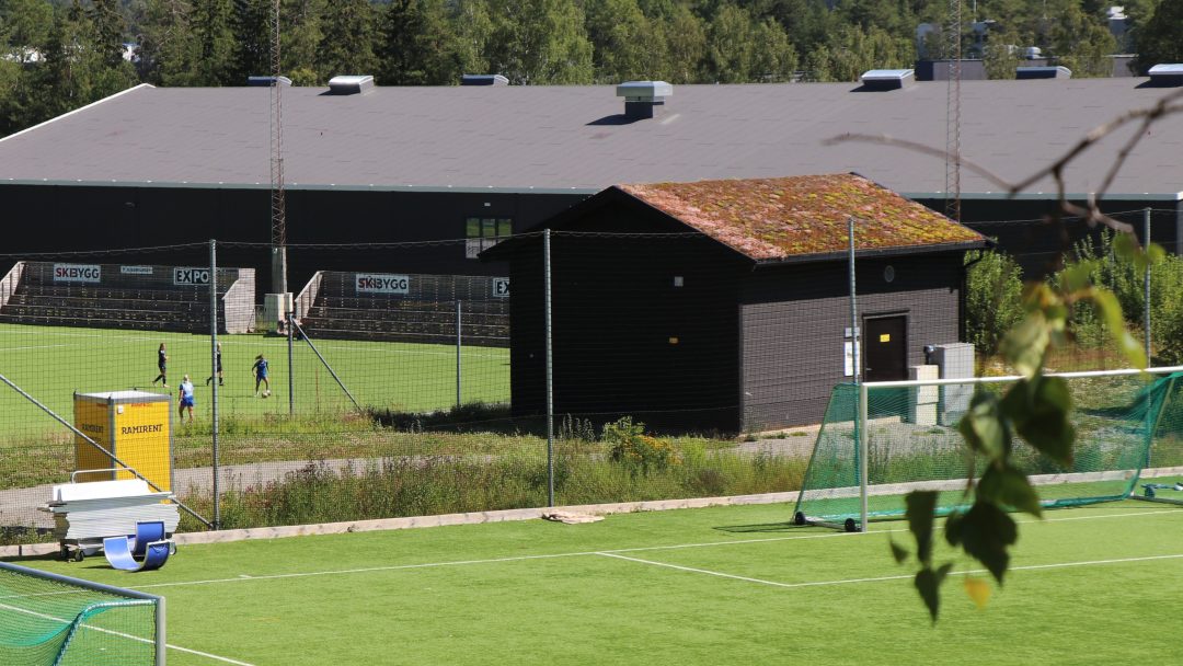 Et lite hus uten vinduer, med torvtak, står mellom to kunstgressbaner