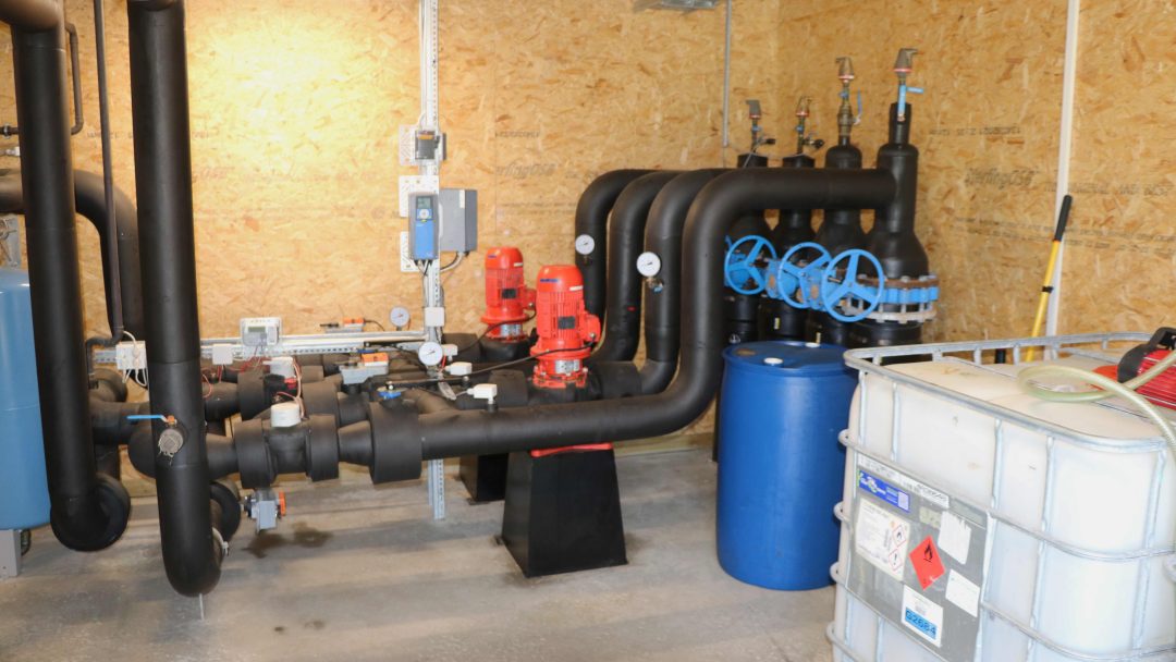 En rekke svarte, store rør forsvinner ned i bakken, inne i et lite bygg uten vinduer