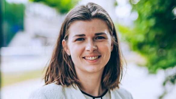 En kvinne med skulderlangt brunt hår smiler, iført en hvit jakke og står utendørs midt i frodig grønt, og legemliggjør energieffektiviseringens ånd.