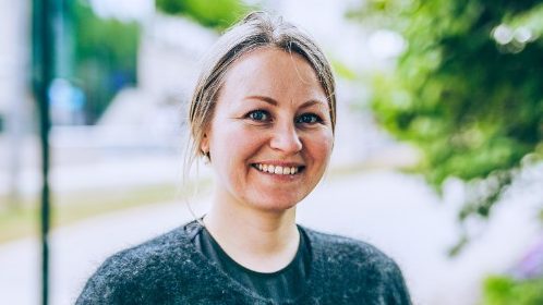 En person i en grå genser smiler utendørs, omgitt av grønne trær, som minner om en skog drevet med selektiv hogst. Den uskarpe urbane bakgrunnen gir en kontrastfull touch til den rolige omgivelsen.