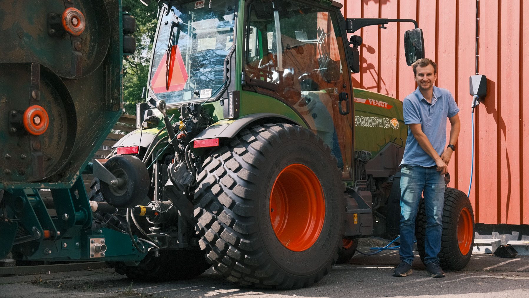 En mann i blå skjorte står ved siden av en grønn traktor med store dekk, parkert ved siden av en rød trebygning i dagslys.