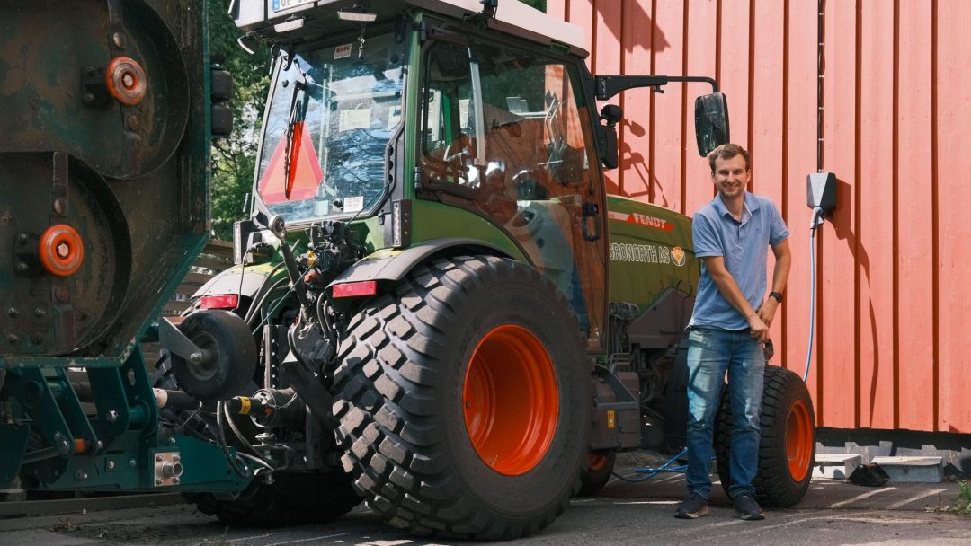 En mann står ved siden av en grønn traktor med store dekk, parkert ved siden av en rød trebygning på en solrik dag.
