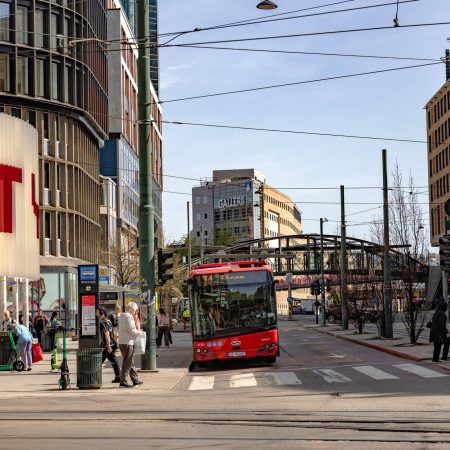 En rød bybuss stopper ved et gatekryss, omsluttet av folk som krysser midt i moderne bygninger mot klar himmel. Scenen gir et dynamisk faggrunnlag for urbant liv, som fanger dansen til hverdagsbevegelser og arkitektonisk bakteppe.