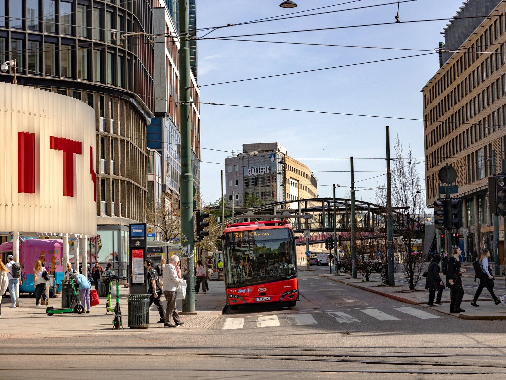 En rød bybuss stopper ved et gatekryss, omsluttet av folk som krysser midt i moderne bygninger mot klar himmel. Scenen gir et dynamisk faggrunnlag for urbant liv, som fanger dansen til hverdagsbevegelser og arkitektonisk bakteppe.